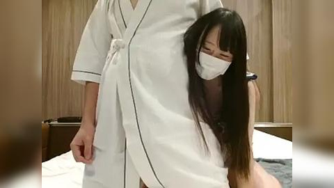 Media: Video of an Asian woman with long black hair, wearing a white robe and face mask, standing in a modern bedroom with wooden walls and a bed.