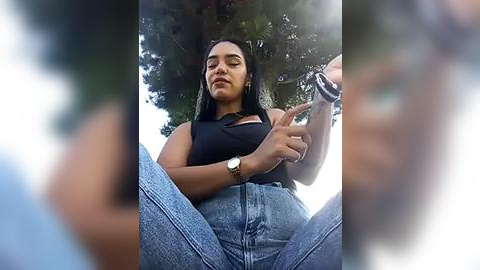 Media: Video of a young woman with medium skin tone and straight black hair, wearing a black top and high-waisted blue jeans, seated outdoors, making a peace sign, with blurred background of trees and sky.