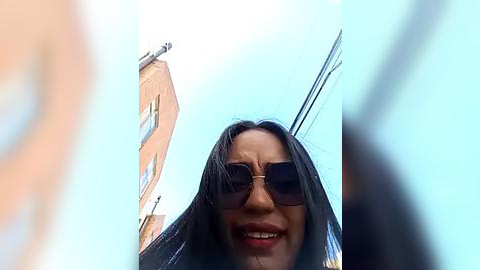 Media: Video of a smiling woman with long black hair, wearing dark sunglasses, standing in front of a beige building under a clear blue sky.