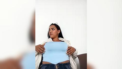 Media: Video of a young woman with medium skin tone, wearing a light blue tank top and jeans, looking contemplative against a plain white background.