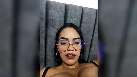 Media: Video of a Latina woman with long black hair, wearing large round glasses and a black bra, against a grey textured backdrop, her lips slightly parted.