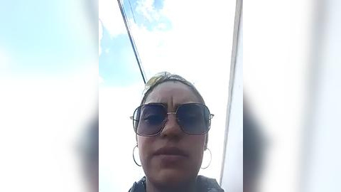 Media: Video of a woman with light brown skin, wearing large aviator sunglasses and a headscarf, standing in front of a white window, with a partly cloudy blue sky in the background.