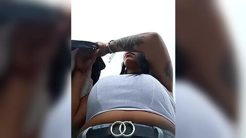 Media: A blurry video shows a tattooed woman wearing a tight, white ribbed crop top and a large, black belt. Her arms are raised, partially obscuring her face, which is turned away.