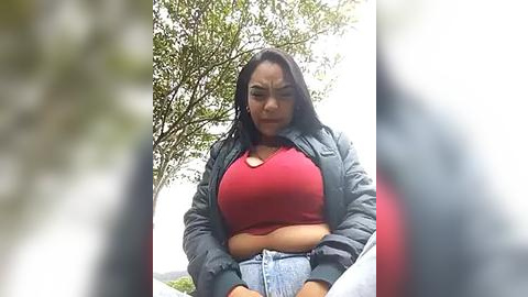 Media: Video of a plus-size woman with dark hair, wearing a red crop top and blue jeans, sitting outdoors among greenery.