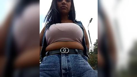 Media: Video of a young woman with long black hair, wearing a pink ribbed crop top and blue high-waisted jeans with a black belt featuring a circular buckle. She stands outdoors with blurred trees and a lamppost in the background.