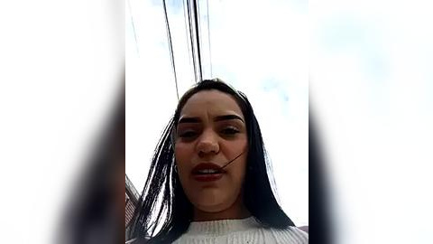 Media: Video of a woman with long black hair, wearing a white sweater, standing in front of a large window with electrical wires, looking slightly annoyed.