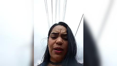 Media: Video of a woman with long black hair, wearing a black choker, her face expressing discomfort as black wires protrude from her forehead, set against a white background.