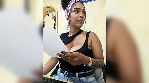 Media: Video of a Latina woman with medium skin tone, medium-sized breasts, wearing a black tank top with a cutout, light-wash jeans, and a purple headscarf. She's seated, holding a piece of paper, in a beige-walled room with a blurred figure in the background.