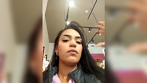 Media: Video of a young Latina woman with medium skin tone and long, straight black hair, styled by a hairstylist. She wears a black jacket and silver earrings. Background shows salon interior with modern lighting and hair styling equipment.