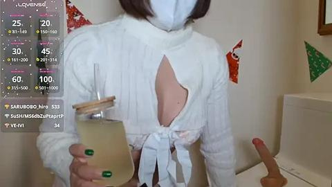 Media: Video of a person wearing a white, sheer, backless top, blue mask, and green nails, holding a glass of yellow liquid. Background shows a white wall with a red and green Christmas decoration.