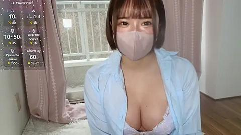 Media: Video of an East Asian woman with short brown hair, wearing a light blue button-up shirt and a pink face mask, revealing cleavage, in a bedroom with pink curtains and wooden floor.