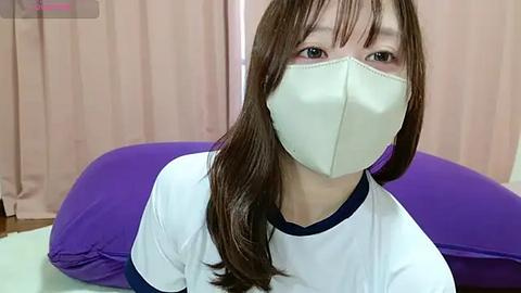 Media: Video of an Asian woman with long brown hair, wearing a white and navy blue t-shirt, a white face mask, and sitting on a purple cushion in a room with beige curtains.