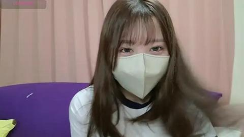 Media: Video of a young woman with long brown hair, wearing a beige face mask, white shirt, and purple pillow. She sits indoors with beige curtains and a yellow piece of paper on a table.