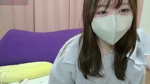 Media: Video of an Asian woman with long brown hair and light skin, wearing a white face mask and a light blue shirt, seated on a purple and yellow bed with a pink curtain backdrop.