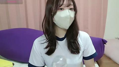 Media: A video of a young woman with long brown hair, wearing a white T-shirt and a white surgical mask, sitting on a purple chair with a beige curtain background.