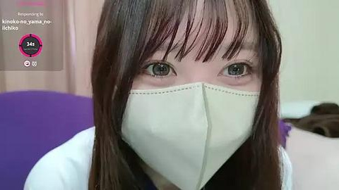 Media: A video of a woman with long brown hair and light skin, wearing a white face mask, green eyes, and a purple top, indoors.