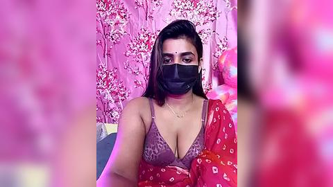 Media: Video of a South Asian woman with medium skin tone, wearing a leopard-print bra, black face mask, and red sari with white floral patterns. Background features pink floral wallpaper.