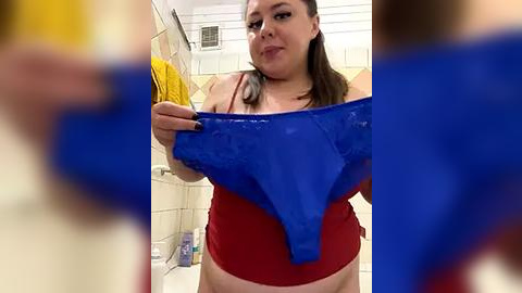 Media: A video of a plus-sized woman in a red top and blue lace bra, holding the bra up in a tiled bathroom with yellow towels and toiletries.