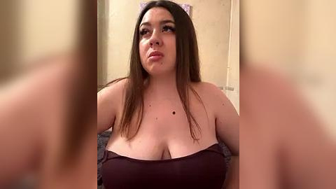 Media: Video of a plus-sized woman with long brown hair, wearing a strapless maroon top, sitting in a dimly lit room with beige walls. She has a neutral expression.