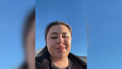 Media: A close-up video of a smiling woman with fair skin, dark hair, and glasses, set against a clear blue sky background. She wears a black jacket and a purple shirt.