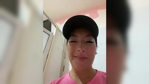 Media: Video of an Asian woman with light skin tone, wearing a pink top and black cap, smiling, standing in a bathroom with white walls and a window.