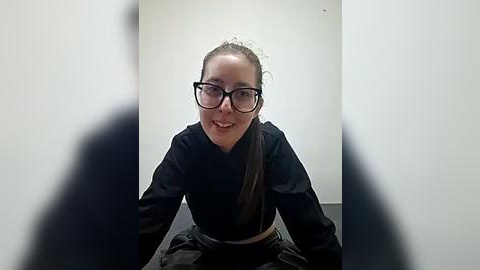 Media: Video of a young woman with glasses, smiling, wearing a black long-sleeve top and a black skirt, seated in a minimalist room with white walls.