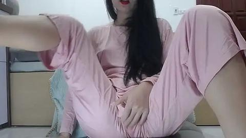 Media: Video of a light-skinned woman with long black hair, wearing pink pajamas, seated on a chair, legs spread, hand on her crotch, in a domestic setting.