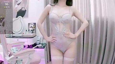 Media: Video of a slim, fair-skinned woman with long black hair, wearing a sheer, light pink lingerie bodysuit with thigh-high stockings. She stands confidently in a modern, well-lit bedroom with a vanity and mirror in the background.