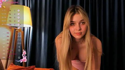 Media: Video of a young blonde woman with long hair, wearing a strapless pink top, leaning forward on a bed, with a calendar and lamp in the background.