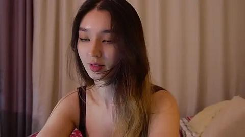 Media: Video of an East Asian woman with long, straight, dark hair, wearing a black spaghetti strap top, sitting in a dimly lit room with beige curtains and a floral pillow.