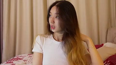 Media: Video of an Asian woman with long, straight, dark hair transitioning to blonde at the ends, wearing a white ribbed top, sitting on a bed with a red patterned duvet and beige curtains in the background.