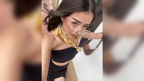Media: A video of a young woman with medium skin tone, long dark hair, and wearing glasses, a black crop top, and a yellow scarf. She has a slender build and is indoors, with blurred figures in the background.