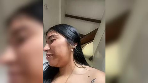 Media: Video of a young Asian woman with straight black hair, medium skin tone, and a small tattoo on her left shoulder. She is seen from the side, looking downward. Background features a white door and a wooden staircase.