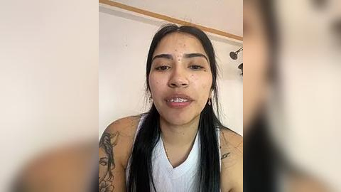 Media: Video of a young Latina woman with long black hair, wearing a white tank top, showcasing tattoos on her arms and neck, with a slightly blurred background.