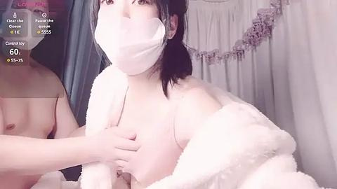 Media: Video of a topless Asian woman with a mask, wearing a fluffy white robe, holding a man's hand, in a dimly lit room.