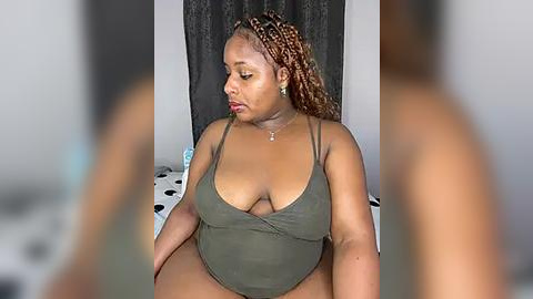 Media: Video of a plus-sized Black woman with medium brown skin, wearing a low-cut olive-green bodysuit, sitting on a bed with polka-dot sheets. Her hair is styled in braids. Background shows a dark curtain.