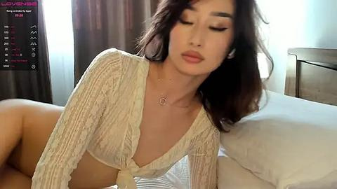 Media: A video of an Asian woman with long dark hair, wearing a sheer, off-shoulder top and minimal makeup, lying on a bed in a softly lit bedroom.