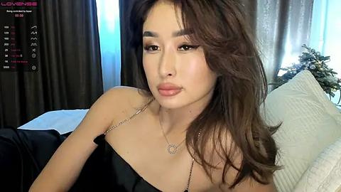 Media: Video of an Asian woman with long brown hair, wearing a black off-shoulder top, sitting on a bed with white pillows. She has a light complexion, makeup, and a delicate necklace. Background shows curtains and a Christmas tree.