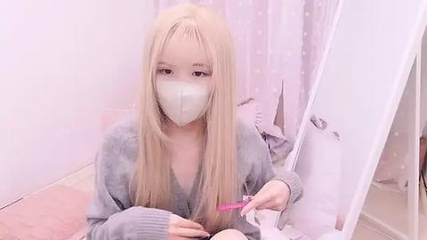 Media: A young Asian woman with long blonde hair and fair skin, wearing a face mask, a gray sweater, and pink nail polish, sits on a pink carpet, holding a pink pen.