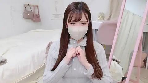 Media: Video of an Asian woman with long brown hair, wearing a white face mask, light gray sweater, and white button-up shirt, sitting on a white bed in a softly lit, minimalistic bedroom.