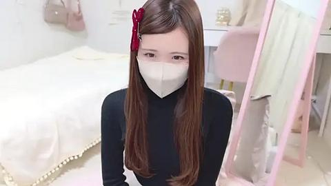 Media: Video of an Asian woman with long brown hair, wearing a black turtleneck, white face mask, and a red flower hairpin, kneeling on a pink carpet in a pastel-colored bedroom with a white bed and pink furniture.