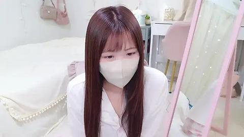 Media: A video of a young Asian woman with long brown hair, wearing a white face mask, sitting on a bed in a minimalist, bright room. She appears to be reading a book.