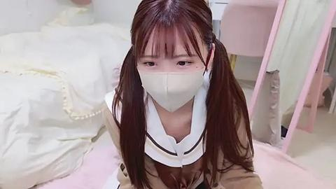 Media: Video of an Asian woman with long, dark hair in pigtails, wearing a white surgical mask and a light brown jacket, sitting on a pink floor in a soft, pastel-toned room with a white bed and pink mirror.