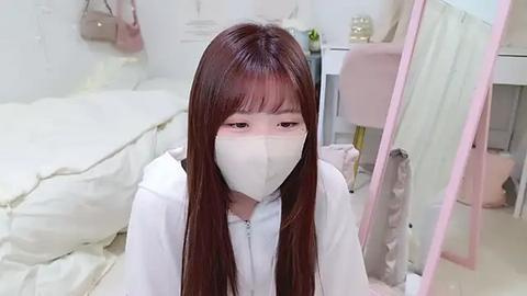 Media: Video of a young Asian woman with long, straight brown hair, wearing a white face mask and hoodie, kneeling in a minimalist, white bedroom with a bed, desk, and mirror in the background.