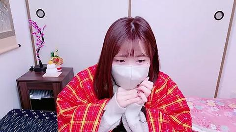 Media: A video of a young Asian woman with straight brown hair, wearing a red plaid jacket and a white mask, seated on a pink bed with a floral duvet, in a room with white walls and a wooden nightstand.