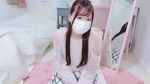 Media: Video of an Asian woman with long brown hair, wearing a white face mask, pink dress, and pink shoes, sitting on a pink blanket in a soft, pastel-toned room.