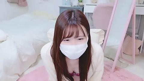 Media: Video of an Asian woman with long brown hair, wearing a white mask, pink cardigan, and red bra, kneeling on a pink rug in a minimalistic, pastel-colored bedroom with white furniture.