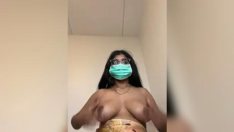 Media: Video of a topless woman with medium-dark skin and long black hair, wearing a turquoise face mask and glasses shaped like frogs. She holds her breasts, revealing a small, light-colored tattoo on her right breast. The background is plain, beige walls.