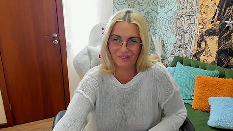 Media: Video of a smiling, blonde, fair-skinned woman with glasses, wearing a gray sweater, sitting on a green sofa with colorful pillows. Background includes a wooden door and patterned tapestry.