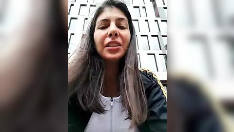 Media: Video of a young woman with long, straight, dark hair, wearing a black jacket with yellow stripes, standing in front of a modern building with a grid-like fa\u00e7ade.